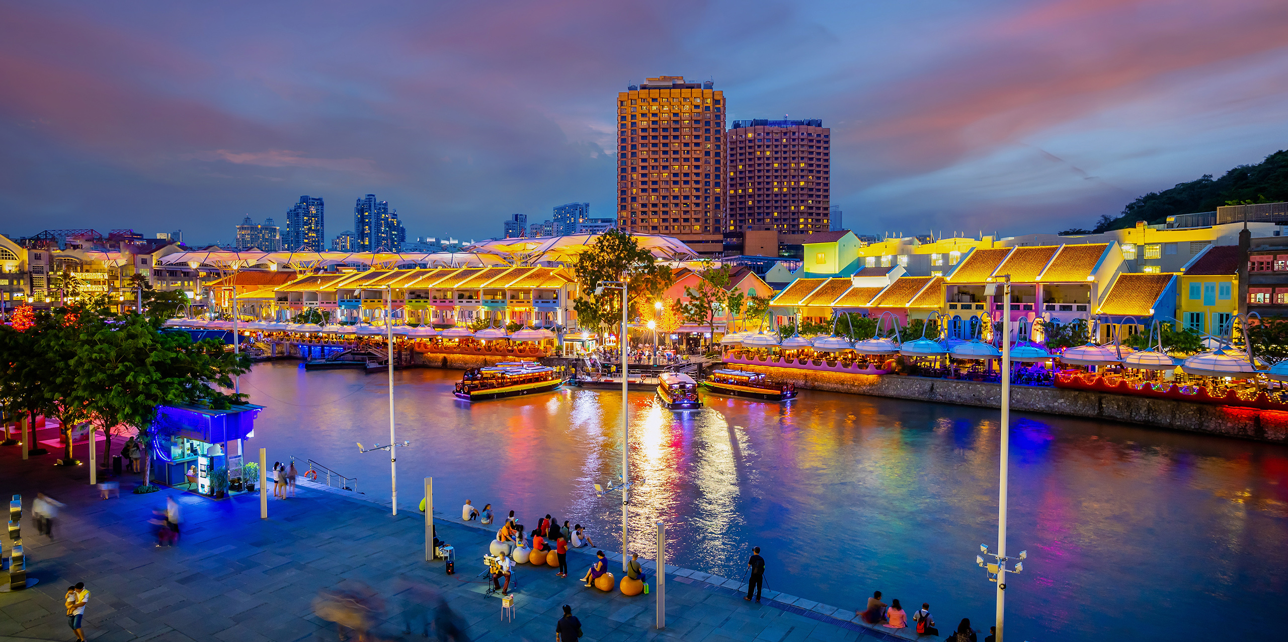 Place page Hero - Clarke Quay Singapore The night lights with vibrant colors in Singapore's Clarke Quay area, with passersby enjoying the riverside walk