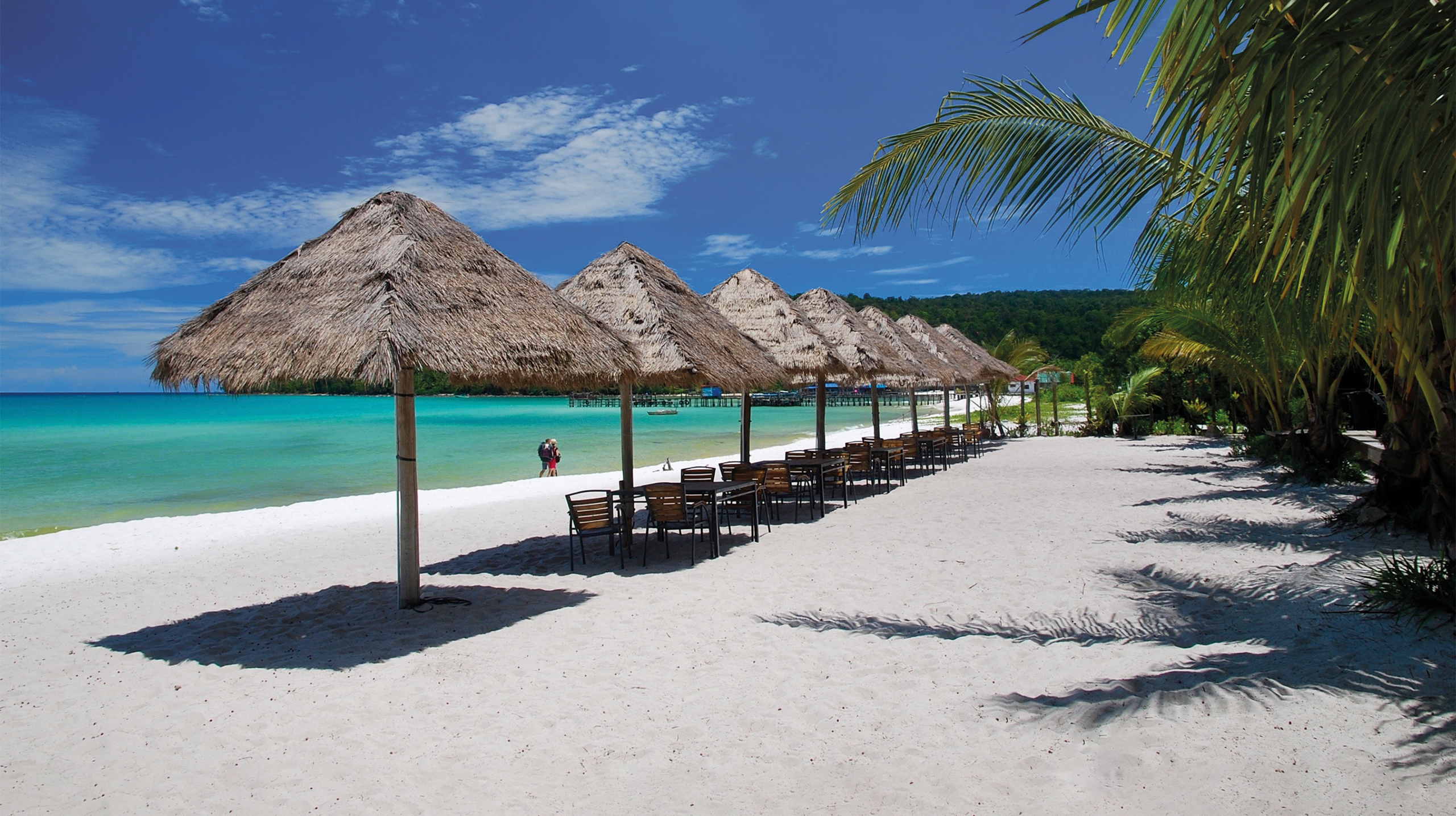 White-sand beach lined with straw-thatched umbrellas next to azure waters in Cambodia's coast.