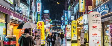 Image of a Korean city in the rain at night, with neon signs and people walkign